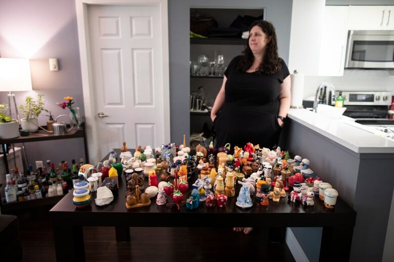 A white woman stands against a wall while dozens of salt and pepper shakers lie on a table in front of her.