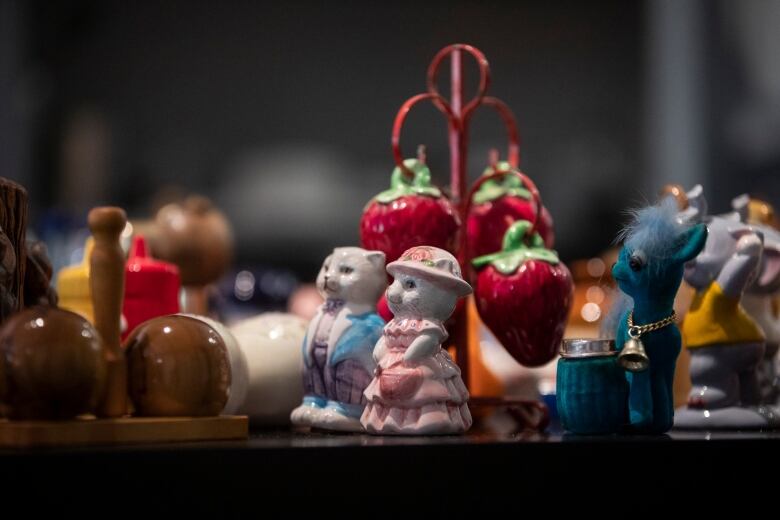 Dozens of salt and pepper shakers lie on a table. In focus are two cats standing upright, dressed like a Victorian man and woman, and shakers shaped like a bunch of strawberries. Behind those is a blue horse with a bell.