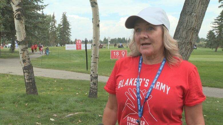 A woman with blond hair wears a red shirt, a blue lanyard, and a white cap at a grassy golf course.