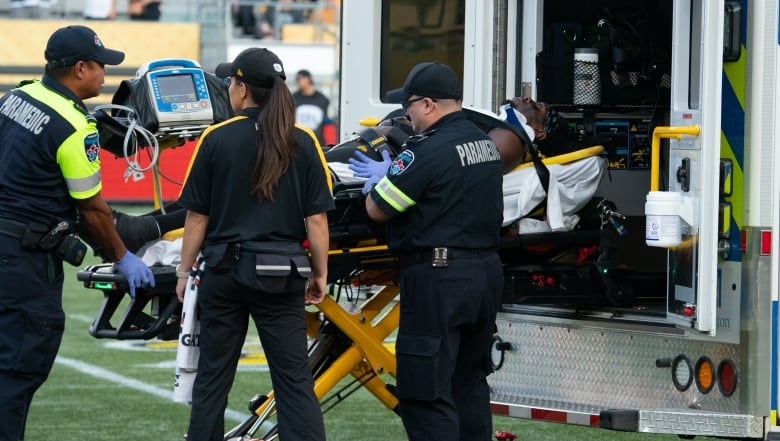 Thee paramedics guide a stretcher, on which lies a Black football player, into an ambulance parked on a football field.