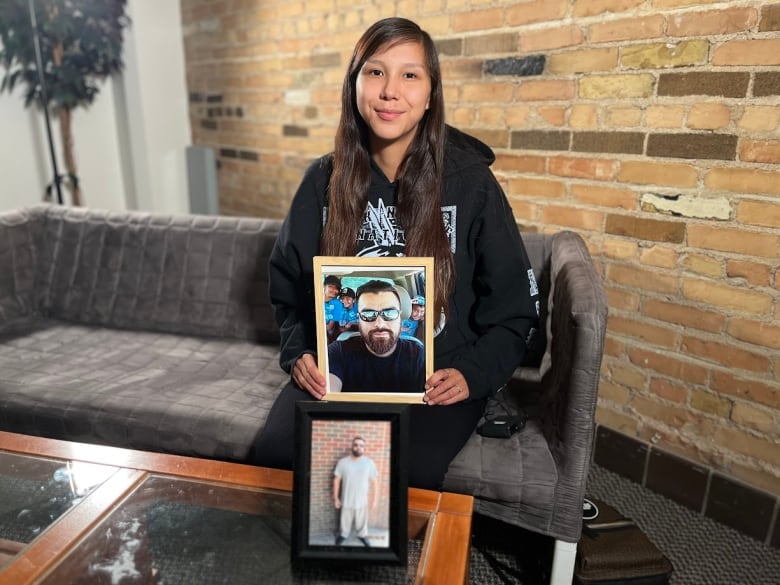 A woman with long black hair sits on a couch and holds a framed photo of a man's face.