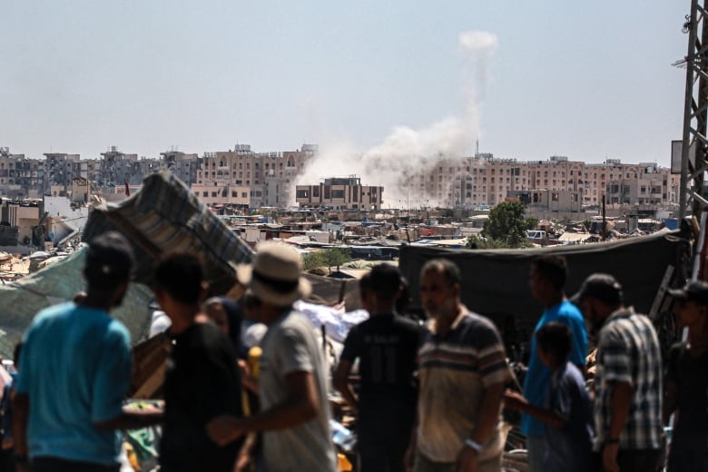 White smoke billows above a city in the background as people in the foreground watch.