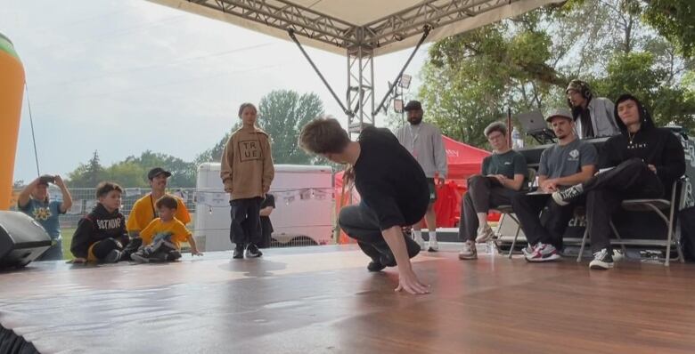 a boy in a t-shirt and sweats dances on the floor of a small stage with a girl looking at him.