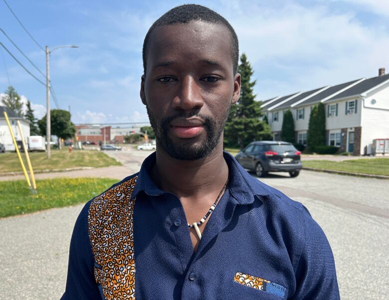 A young man in a dark blue shirt with an orange and white piece of fabric down the right lapel stands in a parking lot. 