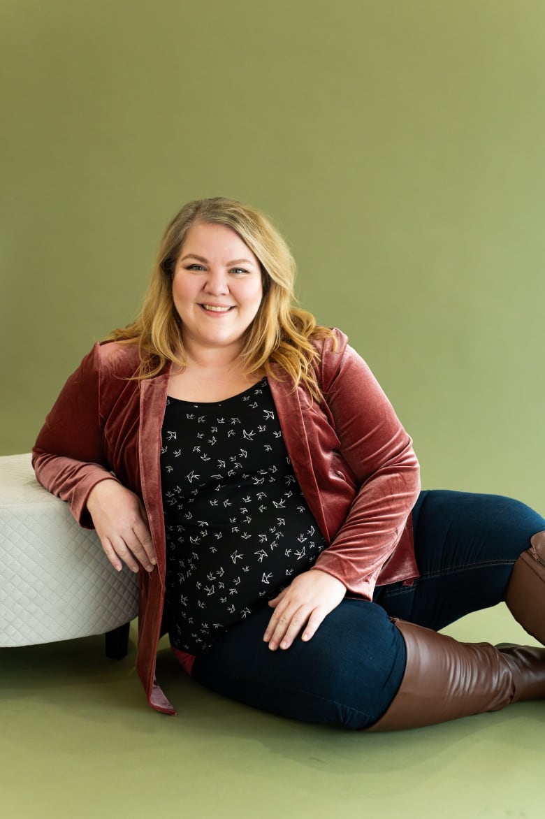 A woman leans on a small couch while sitting on a green backdrop. 