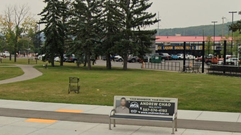 A bus stop bench is seen in the foreground and behind it is a small park with a pathway winding through it to shops in the distance.