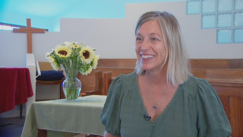 A woman with blond hair smiles in front of a cross in a church.