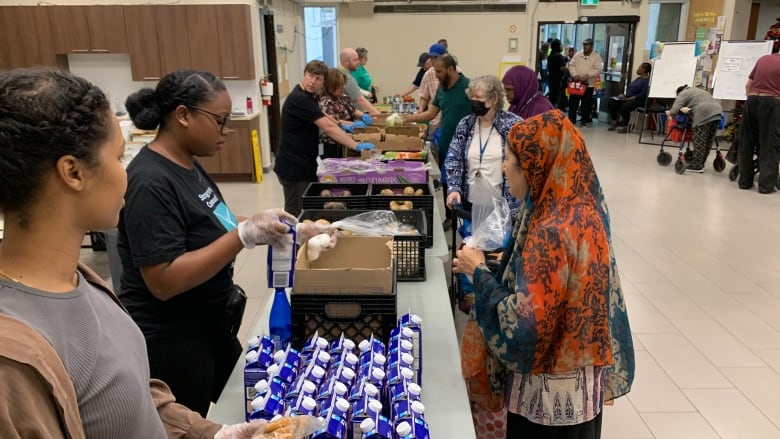 Crates of fresh produce and milk sit on fold out tables in a line. On the left side, volunteers hand out the food to food program recipients taking the food. 