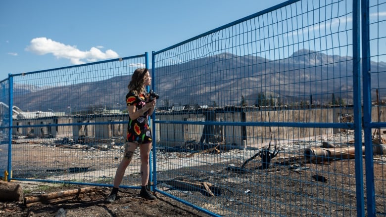 A woman stands before a field of wreckage. The mountains are visible in the background. 