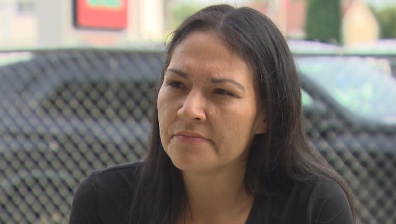 A woman with long black hair, wearing a black t-shirt, looks forward.