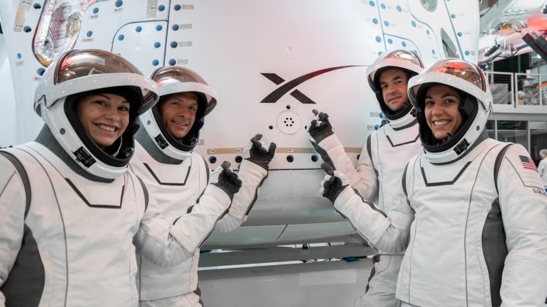 Four astronauts in white spacesuits stand in front of of a SpaceX capsule smiling, pointing at SpaceX logo.