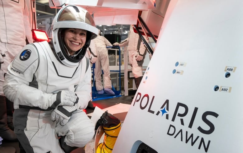 A smiling woman in a white spacesuit kneels in front of a white SpaceX capsule that has the words Polaris Dawn on it.