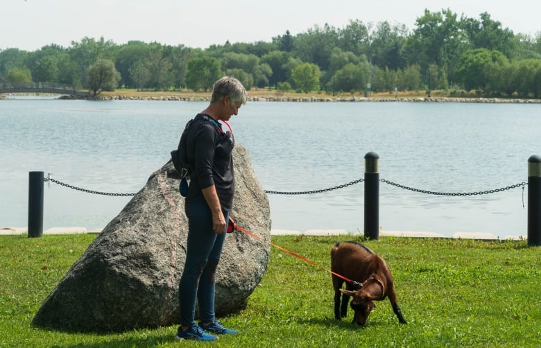 A woman in a black top and blue leggings holds an orange leash attached to a brown goat. Both standing on grass with a body of water in the background