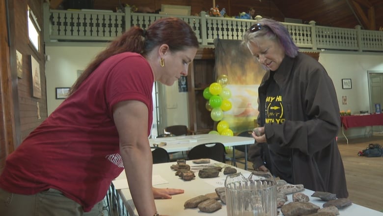 A woman in a red T-shirt stands over a table holding several hand-sized blocks of red sandstone, while another woman looks at a sample in her hand.