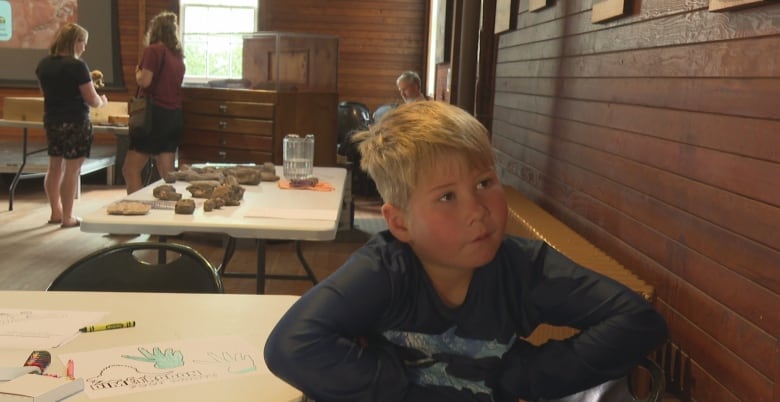 Young boy with blond hair sits at table while people look at trays of fossils behind him.