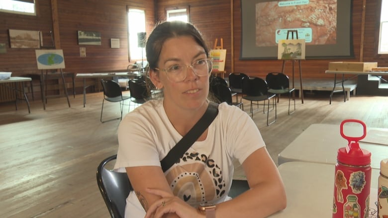 A woman with a white T-shirt and glasses sits at a table in a large room with wooden walls and floors.