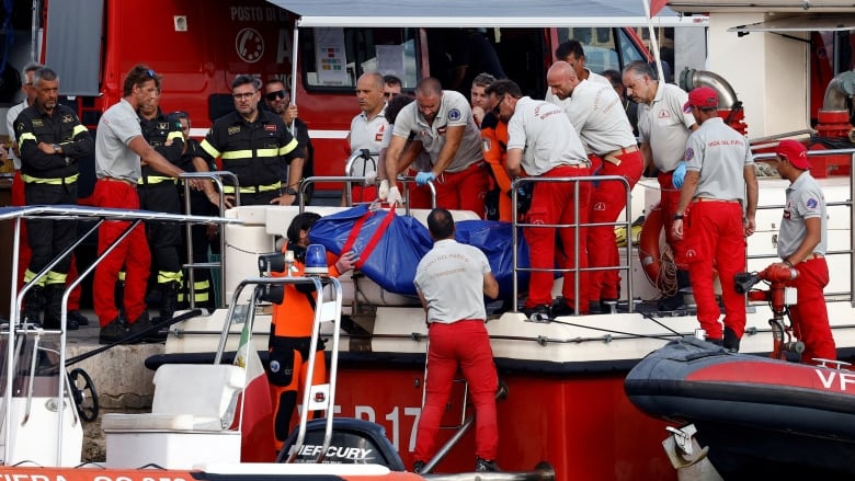 More than a dozen rescue personnel move a blue body bag from a small boat into a larger boat.