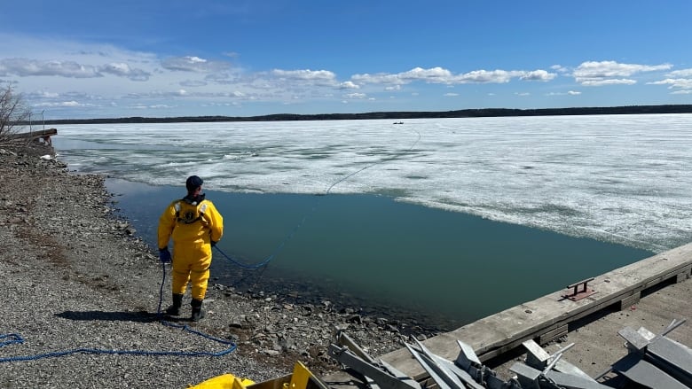 A person in a yellow suit standing at the edge of lake covered in melting ice. He's holding what appears to be a rope that stretches out on to the ice.