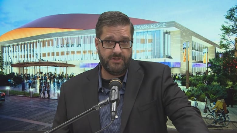 a man in a suit jacket stands in front of a screen with a event centre image 