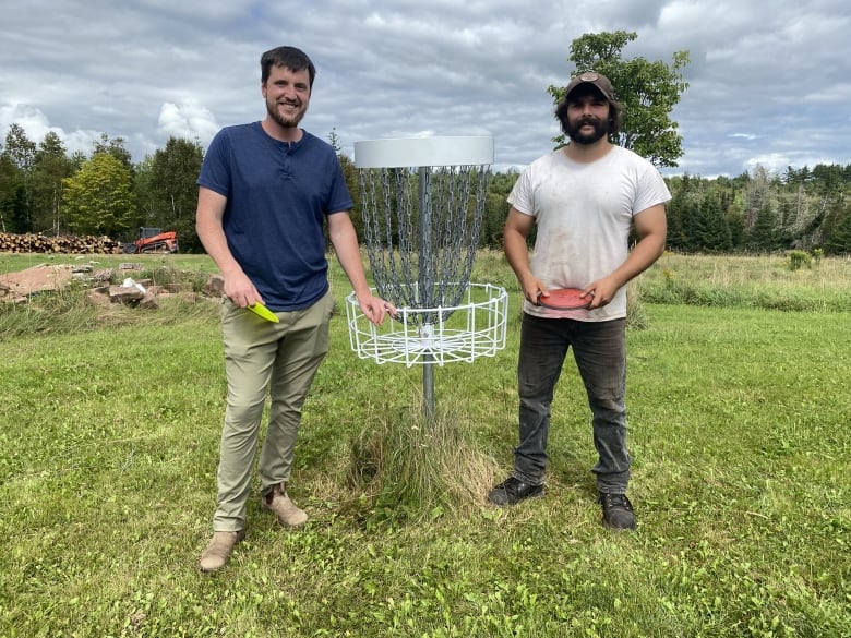 Two men stand beside a disc golf basket holding discs.