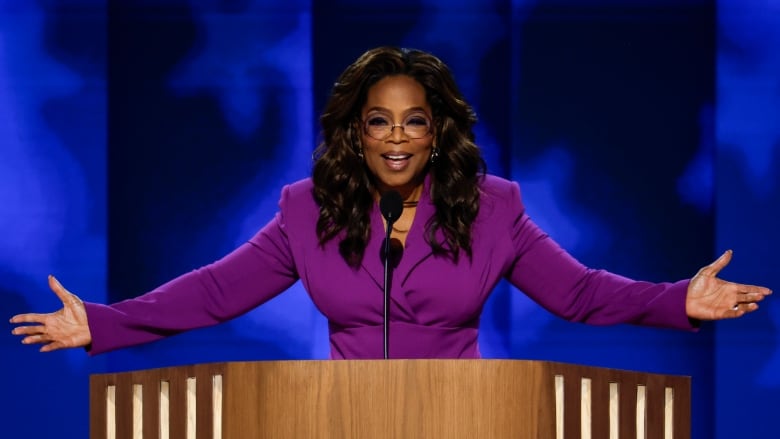 A woman in a purple suit at a podium smiles in front of a blue background. 