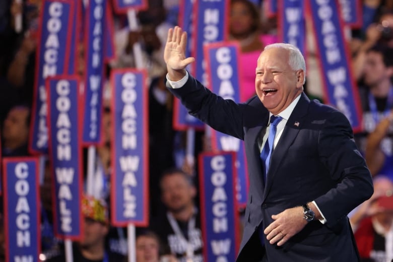 Man waves from stage, with background of people holding up picket signs saying, 