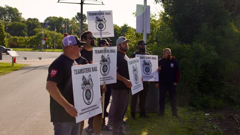 People in matching T-shirts holding 'Teamsters Rail' picket signs stand by a fence off a park path. 