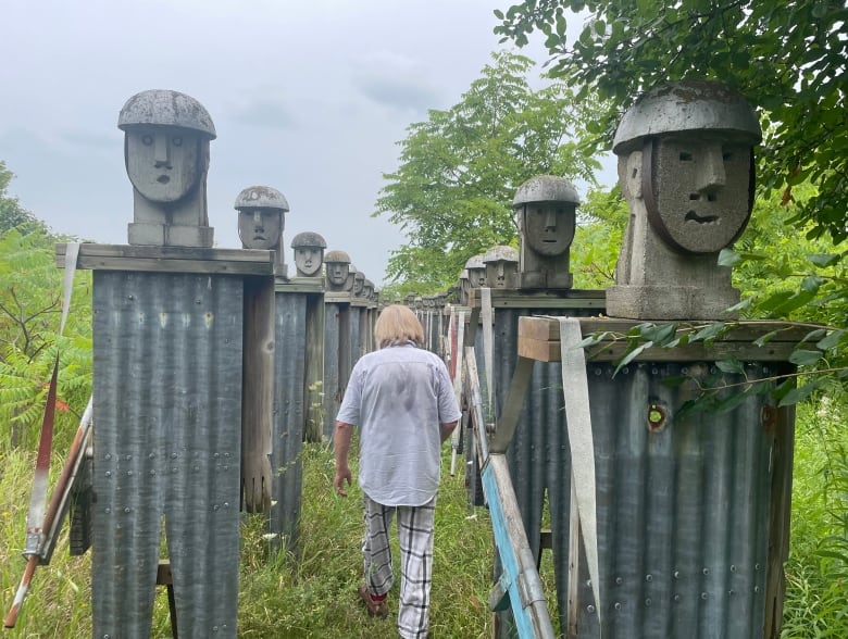 A man with long blonde hair walks between two rows of sculptural figures made from cement and steel to look like soldiers.