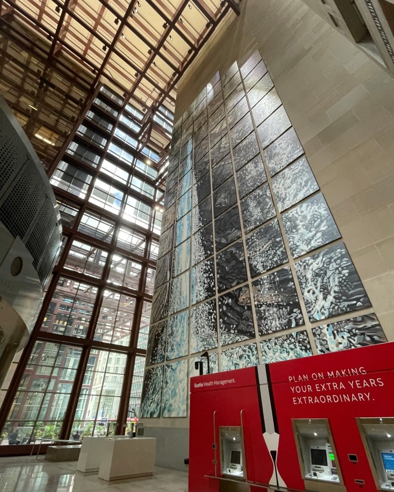 Photo of a multi-story painting of a waterfall. The image is pieced together on 69 individual panels. It hangs inside the atrium of a bank tower, the Scotia Plaza in downtown Toronto. A red ATM blocks the bottom panels of the mural.