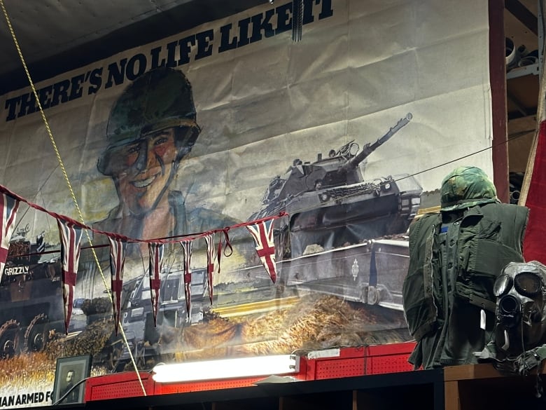 A poster with a drawing of a soldier and tanks. A green army vest and helmet is displayed next to a gas mask.