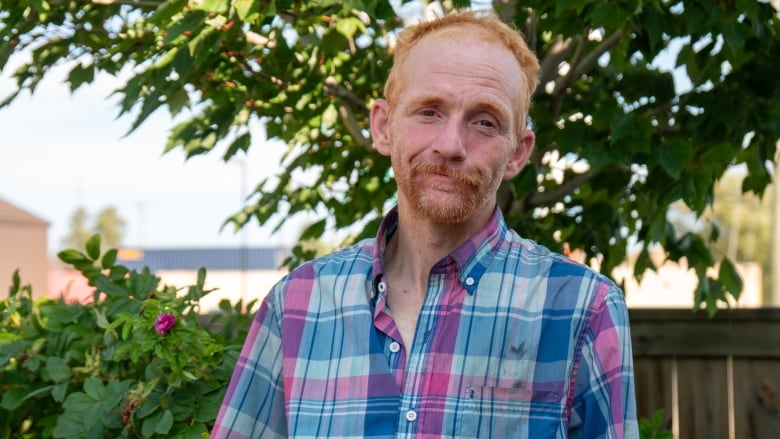 A person wearing a pink and blue plaid shirt stands in front of a bush outside.