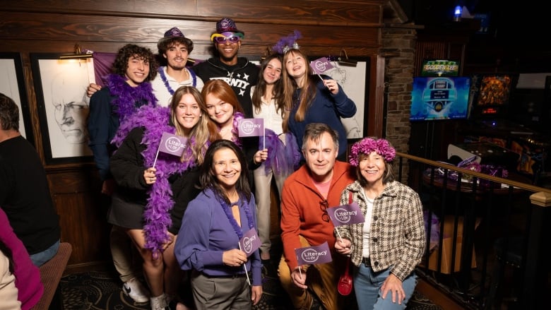 A group of individuals pose in pub, while holding various purple props like a boa, hats, headbands and mini flags.