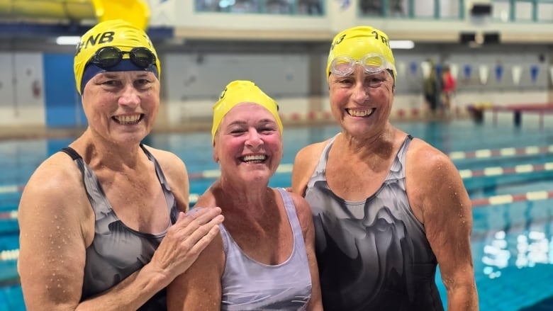 Three older women standing together in swimming suits smiling at camera with pool behind them. 