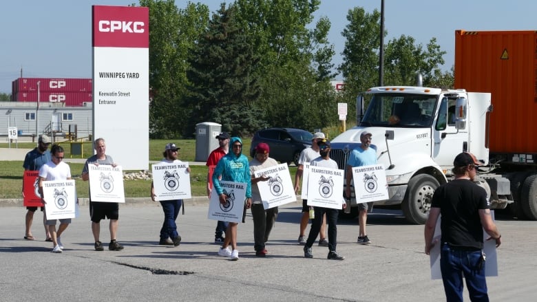 A group of people walk a picket line. A sign for the company, CPKC, stands behind them.