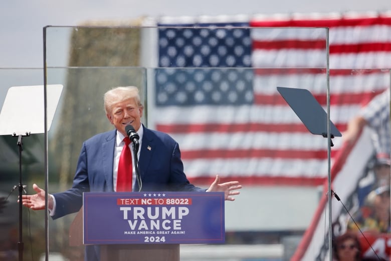 A man in a blue suit and long red tie speaks from behind bulletproof glass during a campaign rally.