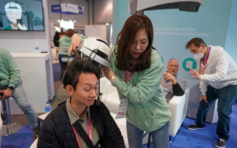 A woman with long dark hair, wearing a green sweater, removes a white helmet from the head of a man with black hair who is wearing a black jacket.