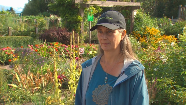 A woman in a blue jacket and cap in a green space filled with flowers and bushes.