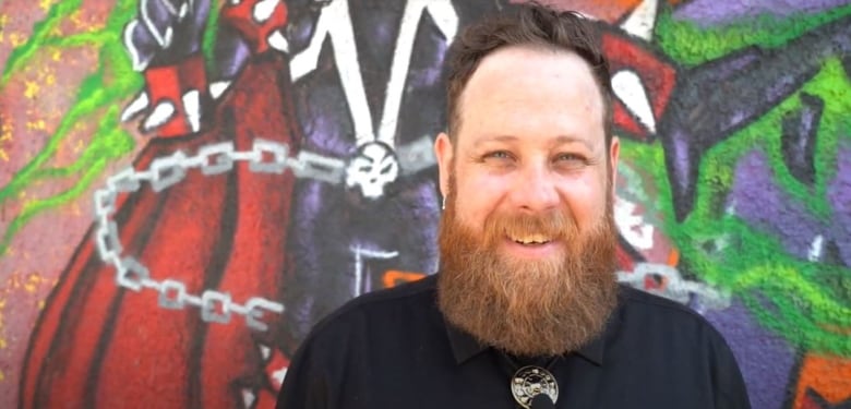 A brunette haired man with a red beard stands smiling in front of a wall of graffiti. He's wearing a black dress shirt and silver bolo tie.