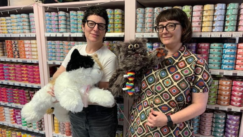 Two women holding stuffed animal cats stand in front of a wall of cat food