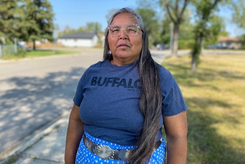 A women with long hair and glasses stands outside while wearing a shirt and skirt with ribbons.