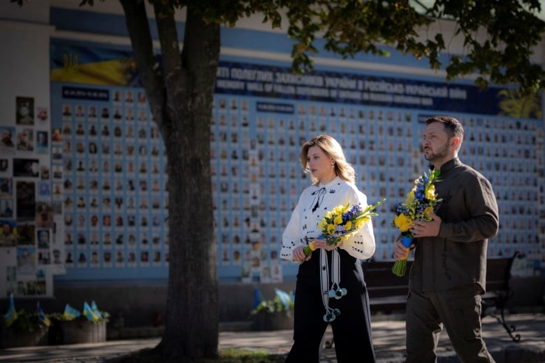 Two people hold flowers in front of a wall of pictures of people.