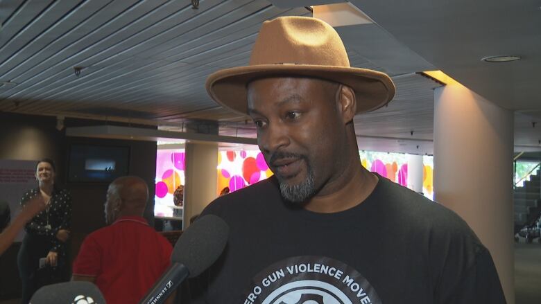 A man with a beard and wide rimmed hat, wearing a Zero Gun Violence Movement T-shirt, speaks to a reporters' microphone inside a theatre lobby. He is visible from the chest up