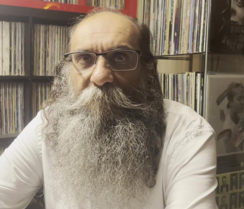 A bald man in glasses with a big bushy beard photographed indoors with stacks of records and books in the shelf behind.