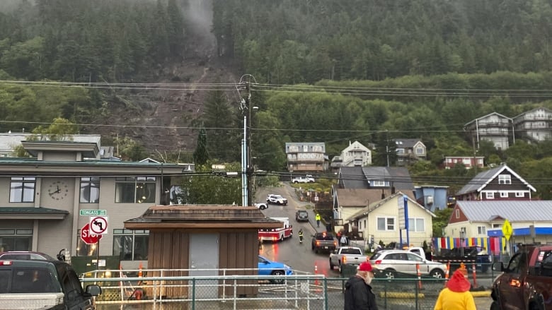 People watch as emergency vehicles move in the background where a large landslide is seen through the trees.