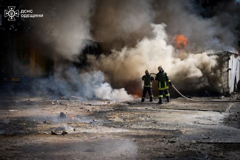 Two firefighters in full uniform and helmet are shown with their backs to the camera utilizing a firehose as massive clouds of smoke and some orange fire are shown.