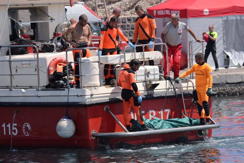 Rescuers dressed in orange bring a body wrapped in a plastic bag aboard a rescue boat. 