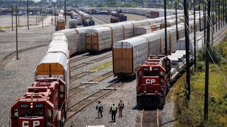 Three railway workers walking between two tracks with stationary railway cars on them, with city of Toronto in the distance. 