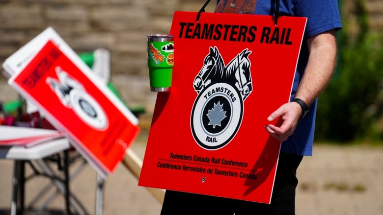 Someone carries a bright red Teamsters Rail sign with the union's name and grey logo.