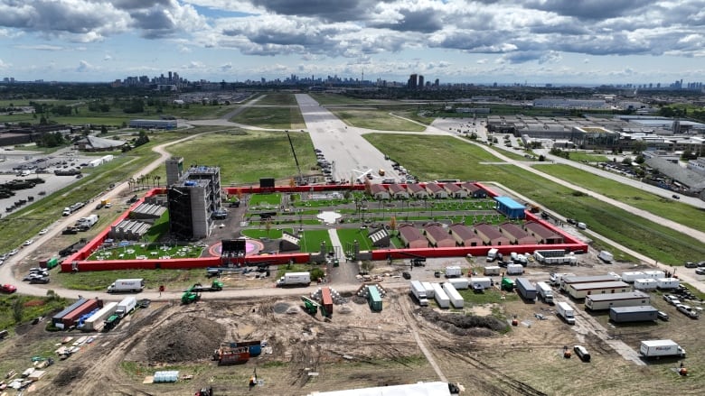 A drone photo shows a small community surrounded by a red wall with a city skyline in the distance. 