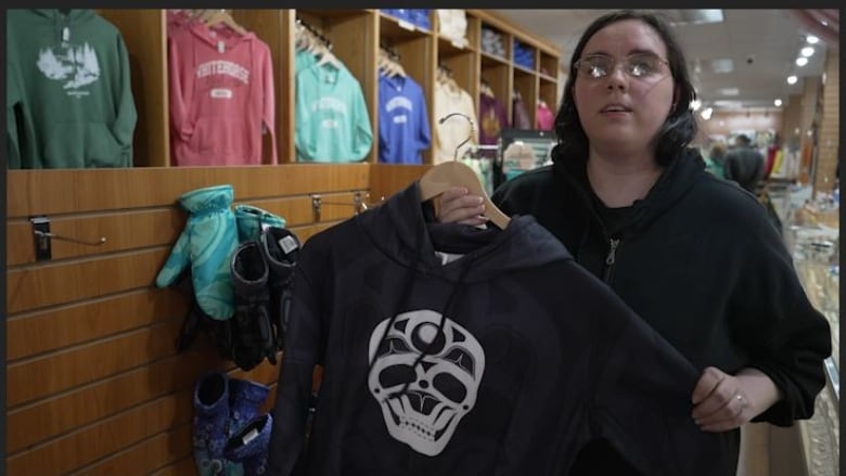 A woman with rounded silver glasses and a black hoodie holds up another black hoodie with a white skull on it.